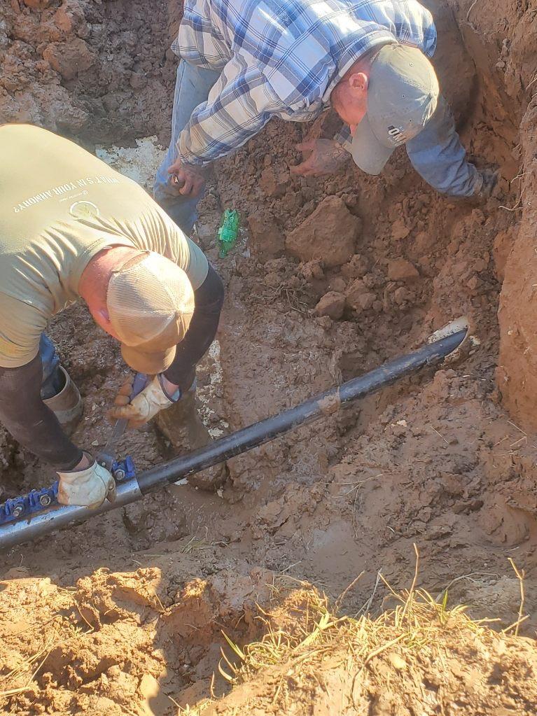 Two men in long-sleeved shirts and baseball caps in a dirt pit, installing a new pipe